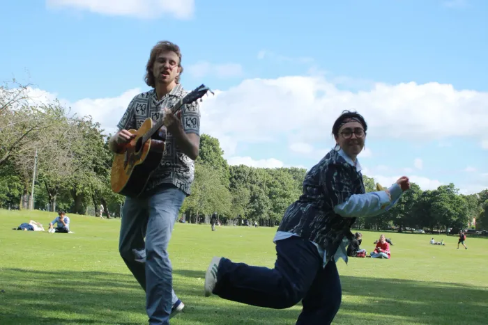 Mick and Bella running across the Meadows under a blue sky, guitar in hand