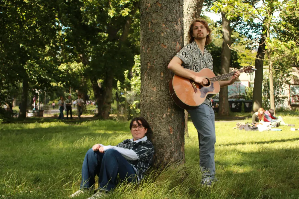 Mick playing acoustic guitar leaning against a tree in the Meadows while Bella sits in the grass below